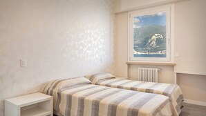 Un dormitorio con dos camas, una mesita de noche blanca, un radiador y una ventana con vistas a la montaña.