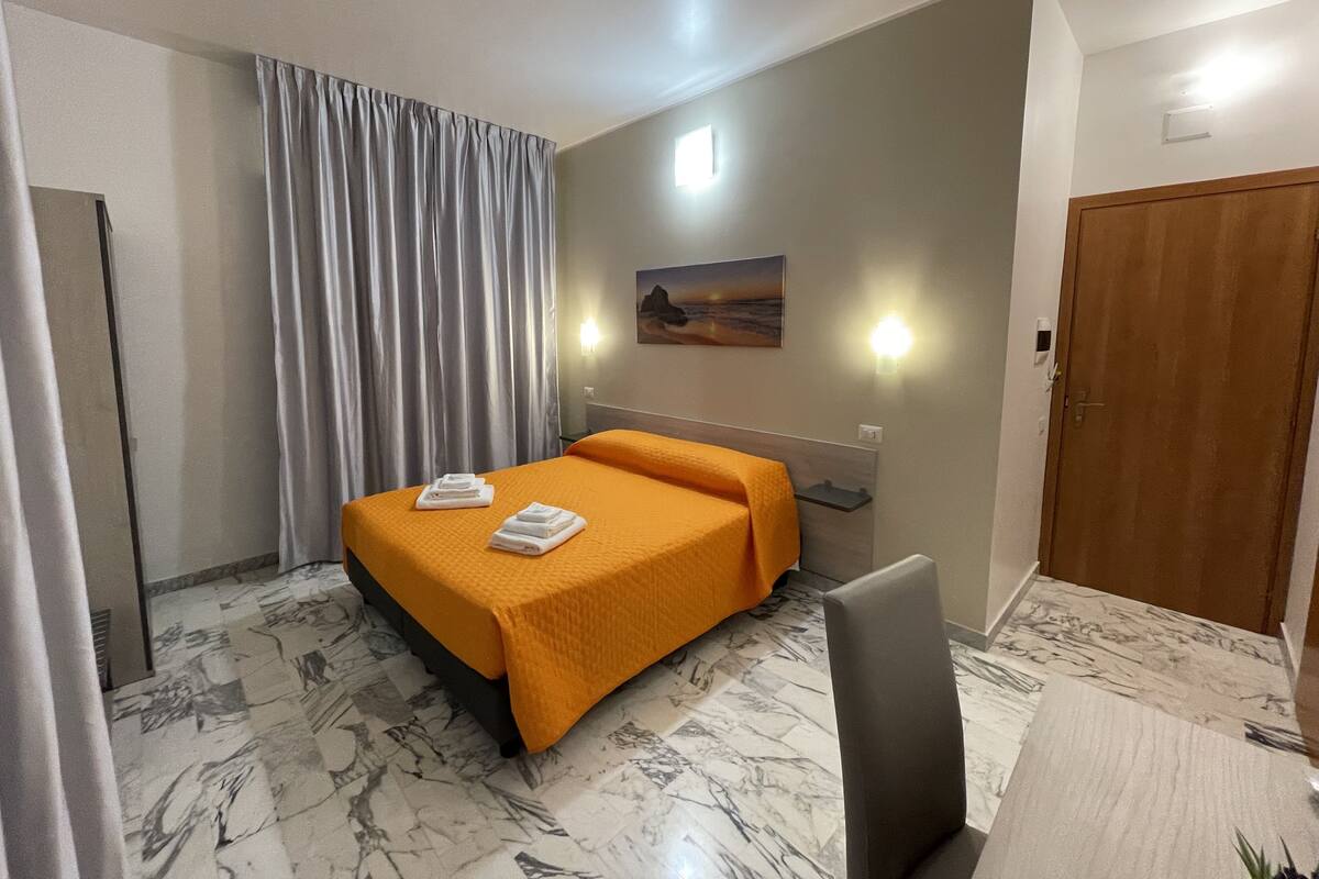 Habitación de hotel con cama, una silla, escritorio, puerta y ventana con cortinas.