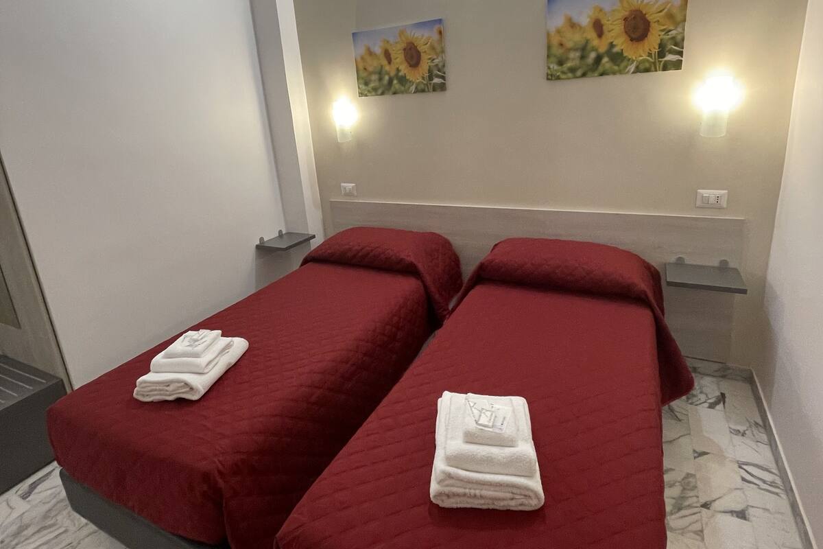 Habitación de hotel con dos camas, cada una con una colcha roja y una toalla blanca doblada. Hay dos apliques de pared y dos cuadros con girasoles en la pared.