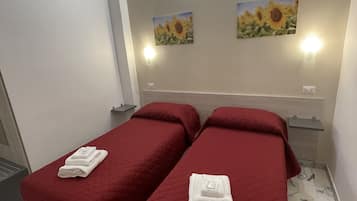 A hotel room with two beds, each with a red bedspread and a folded white towel. There are two wall-mounted light fixtures and two framed pictures of sunflowers on the wall.