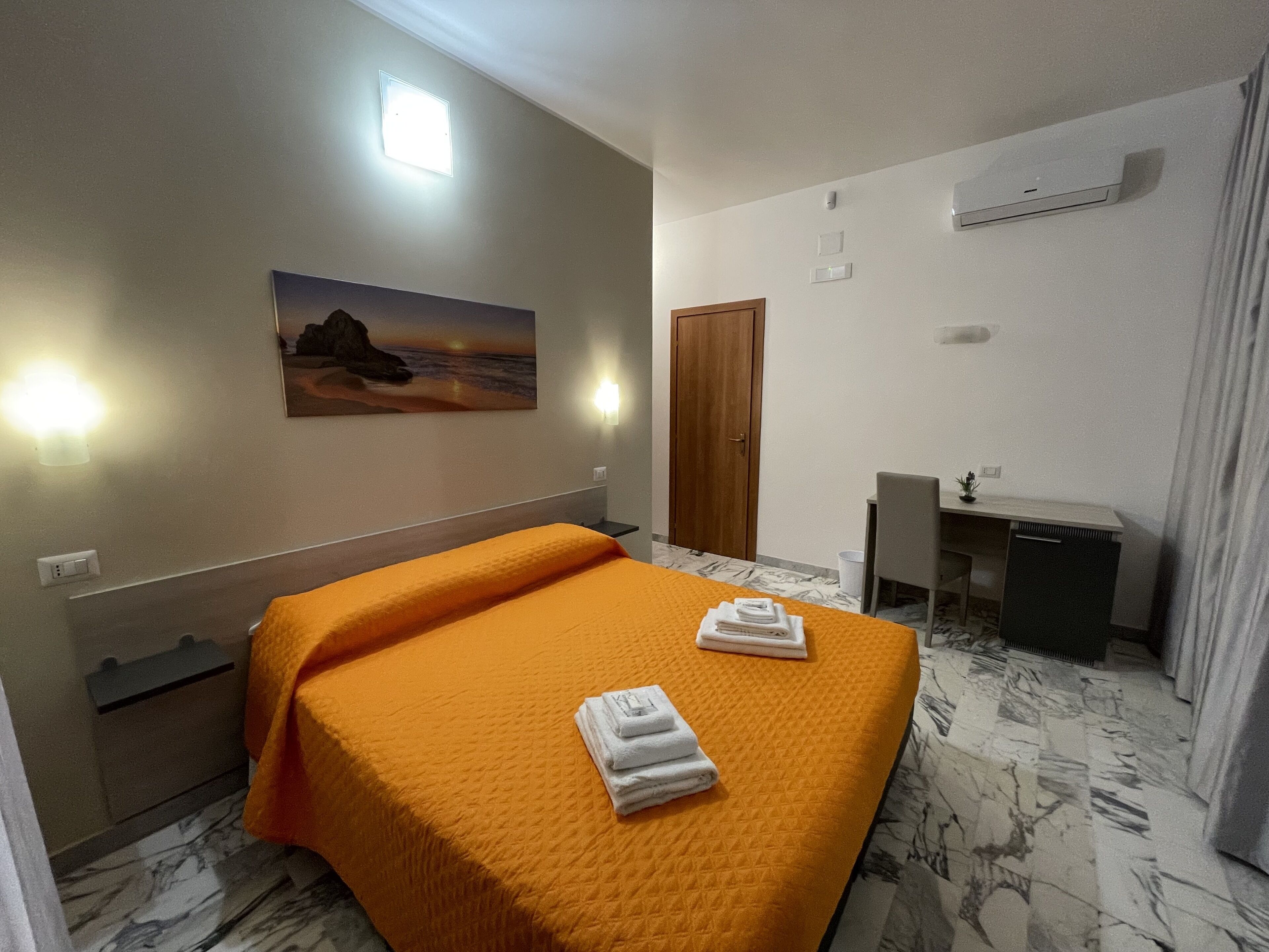 A hotel room with a bed, a desk, a chair, a painting, and an air conditioning unit.