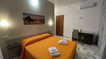 A hotel room with a bed, a desk, a chair, a painting, and an air conditioning unit.