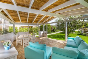 A covered patio with stone flooring, white furniture, and a view of a garden and pool.