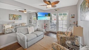A living room with a grey sofa, patterned armchairs, a ceiling fan, a flat-screen TV, and a large window with shutters. - A Fish Bowl at The Truman Annex in Key West (Key West)