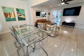 A modern living room with a glass dining table, a brown leather sofa, and a flat-screen TV mounted on the wall. - 6/6 for 12+ Big Heated Pool w/ Tiki Bar-BBQ-Beach (Hollywood)