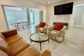 A living room with a sofa, armchairs, a glass coffee table, and a TV mounted on the wall.