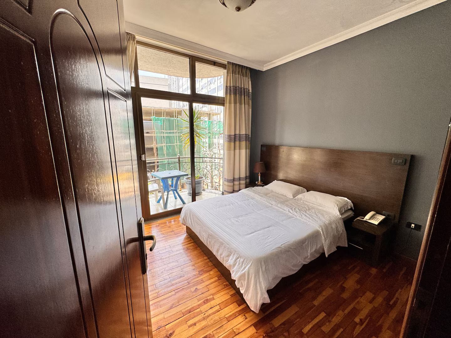 A hotel room with a large bed, a wooden floor, a nightstand with a phone, and a view of a balcony with a table and chair.