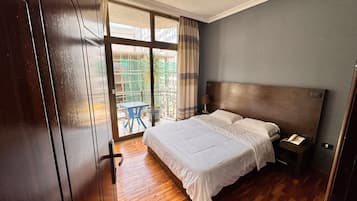 A hotel room with a large bed, a wooden floor, a nightstand with a phone, and a view of a balcony with a table and chair.