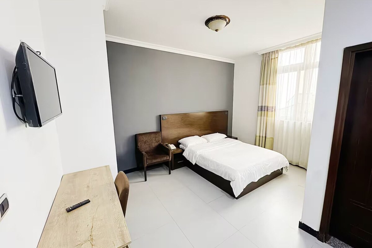 Une chambre d’hôtel avec un lit, un bureau, une chaise, une télévision et une fenêtre avec des rideaux.