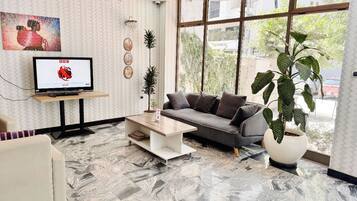A modern living room with a sofa, coffee table, and a TV on a stand. There are plants and a large window providing a view outside.