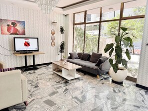 A modern living room with a sofa, coffee table, and a TV on a stand. There are plants and a large window providing a view outside. - Rankset Guesthouse apartment (Addis Ababa)