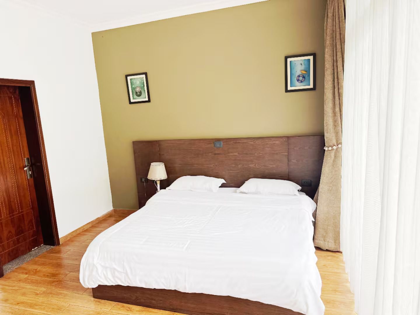 Habitación de hotel con cabecera de madera, cama con ropa blanca, dos cuadros enmarcados en la pared, una lámpara de noche y una puerta.