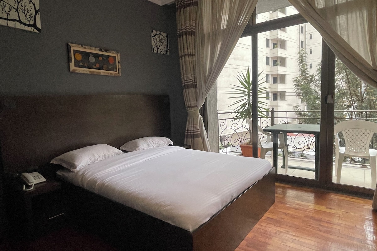 A hotel room with a large bed, a telephone, and a balcony with a view of buildings and a plant.