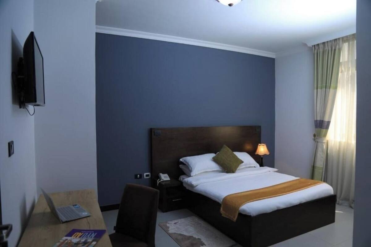 A hotel room with a bed, a desk with a laptop, a chair, a nightstand with a lamp, and a TV on the wall.