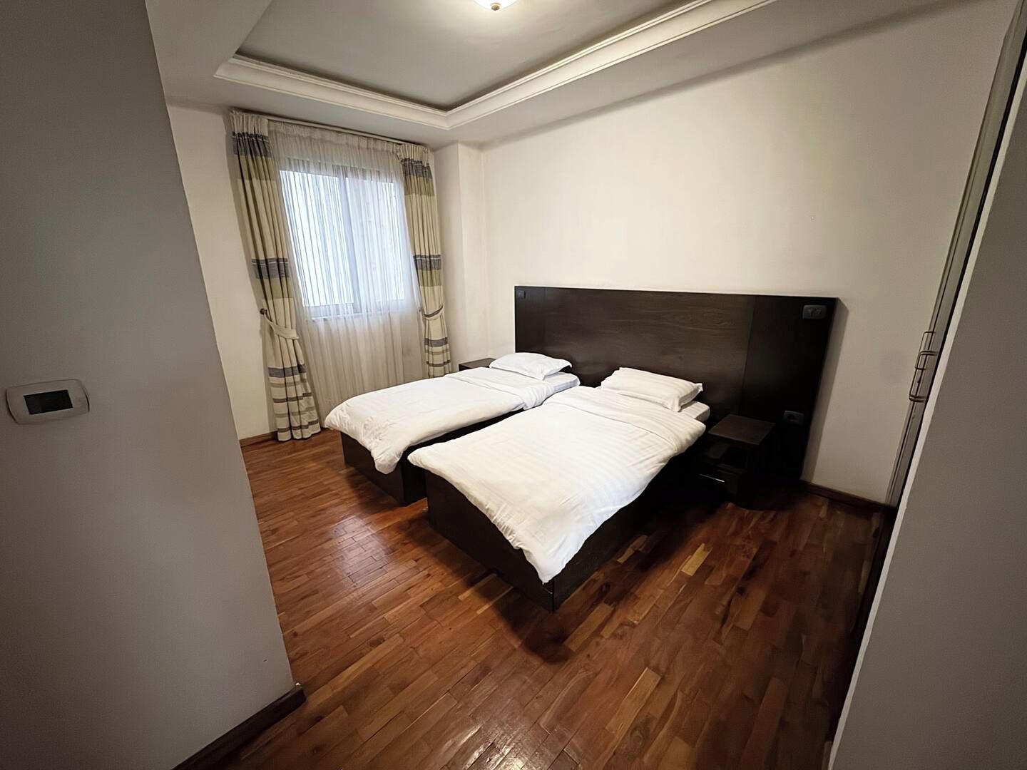 A hotel room with two beds, wooden flooring, a dark headboard, and a window with curtains.