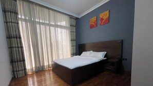 Un dormitorio con piso de madera, una cama con ropa blanca, una mesita de noche, un televisor montado en la pared y una ventana con cortinas translúcidas.