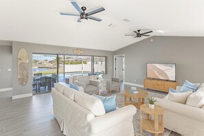 A spacious living room with a large sofa, a coffee table, and a TV. There is a ceiling fan and a sign that reads "Beach House".