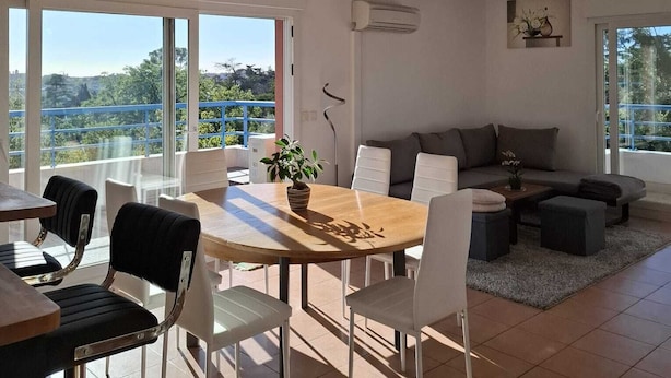 A modern living room with a dining area, large windows, and a view of greenery.