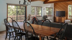 A spacious dining area with a wooden table and chairs, large windows offering views of trees, and a television on a wooden cabinet. - Lay-Z-Bones ~ AC & Pool Access! (Dorrington)
