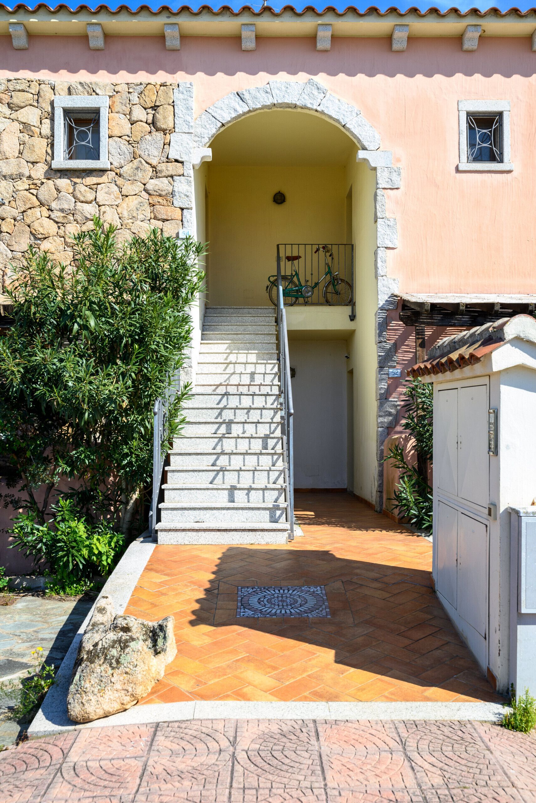 Eine Steintreppe führt zu einer Tür, einer gefliesten Terrasse und einem kleinen Garten mit einem Stein und Pflanzen.