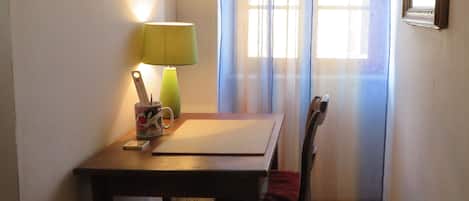 Une petite pièce avec une table en bois, une chaise, une lampe et une fenêtre avec des rideaux.