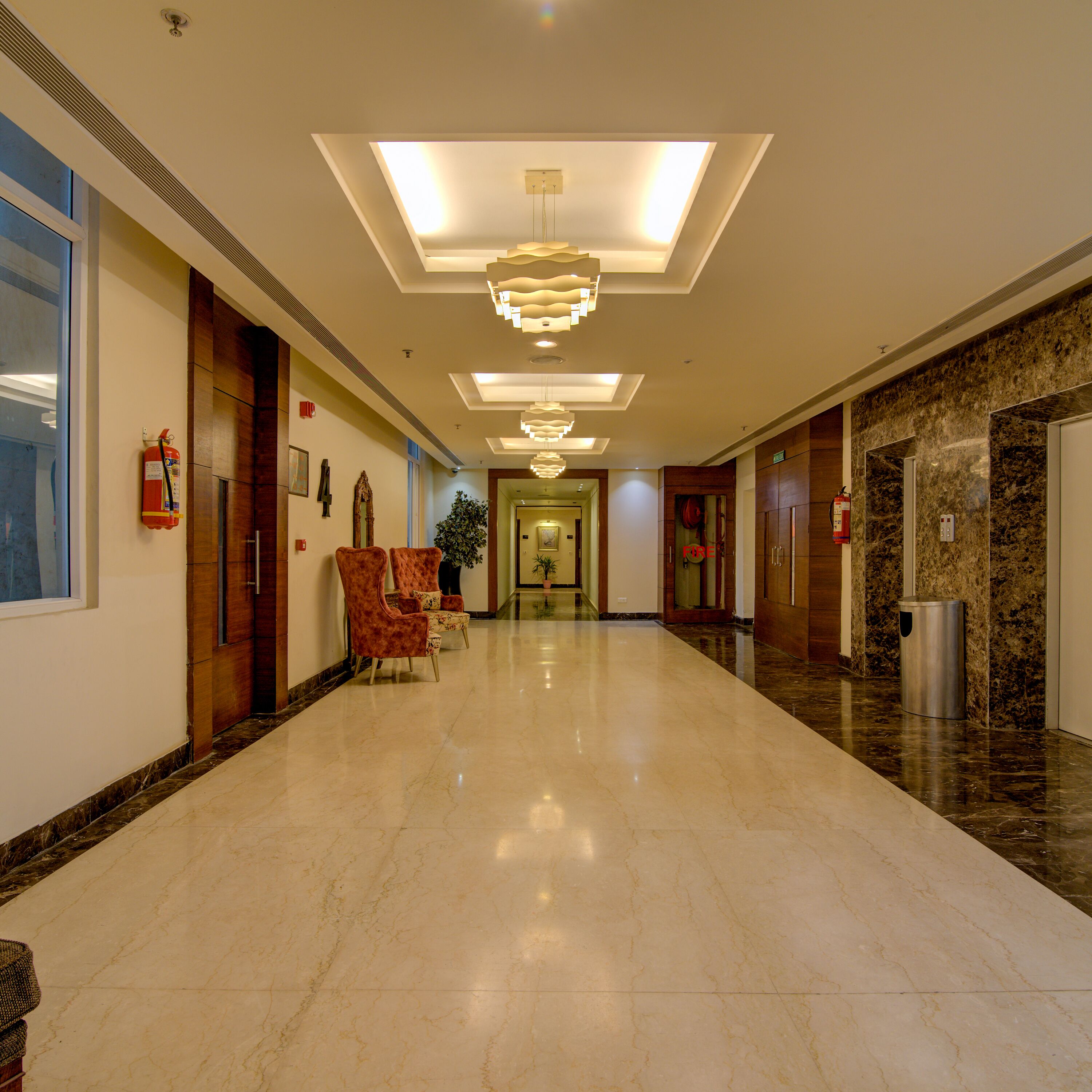 Un vestíbulo de hotel con suelo de mármol, un extintor, una planta y una escalera que sube a un nivel superior.