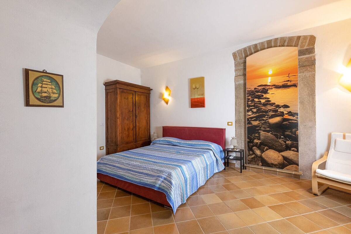 Quarto com cama, guarda-roupa de madeira, mesa de cabeceira, abajur e parede com pintura de pôr do sol.