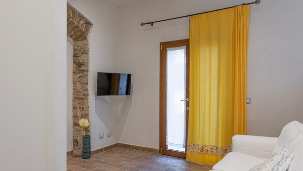 A living room with a white sofa, a television mounted on the wall, and a yellow curtain covering a sliding glass door. - Agriturismo San Giuseppe (Piombino)