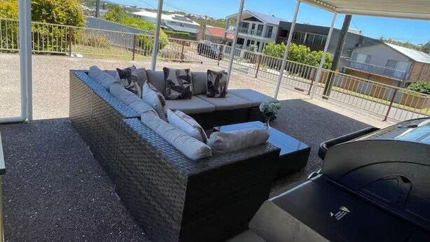 A covered outdoor seating area with a sofa and cushions, a car, and a view of houses and greenery. - Cheerful beach house at redhead beach (Redhead)