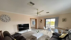 A living room with a flat-screen TV, dark sofas, a coffee table, and a dining area with chairs and a table. - Cheerful beach house at redhead beach (Redhead)