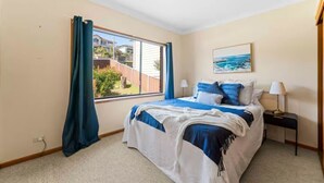 A bedroom with a bed, a window with curtains, a nightstand, and a painting on the wall. - Cheerful beach house at redhead beach (Redhead)