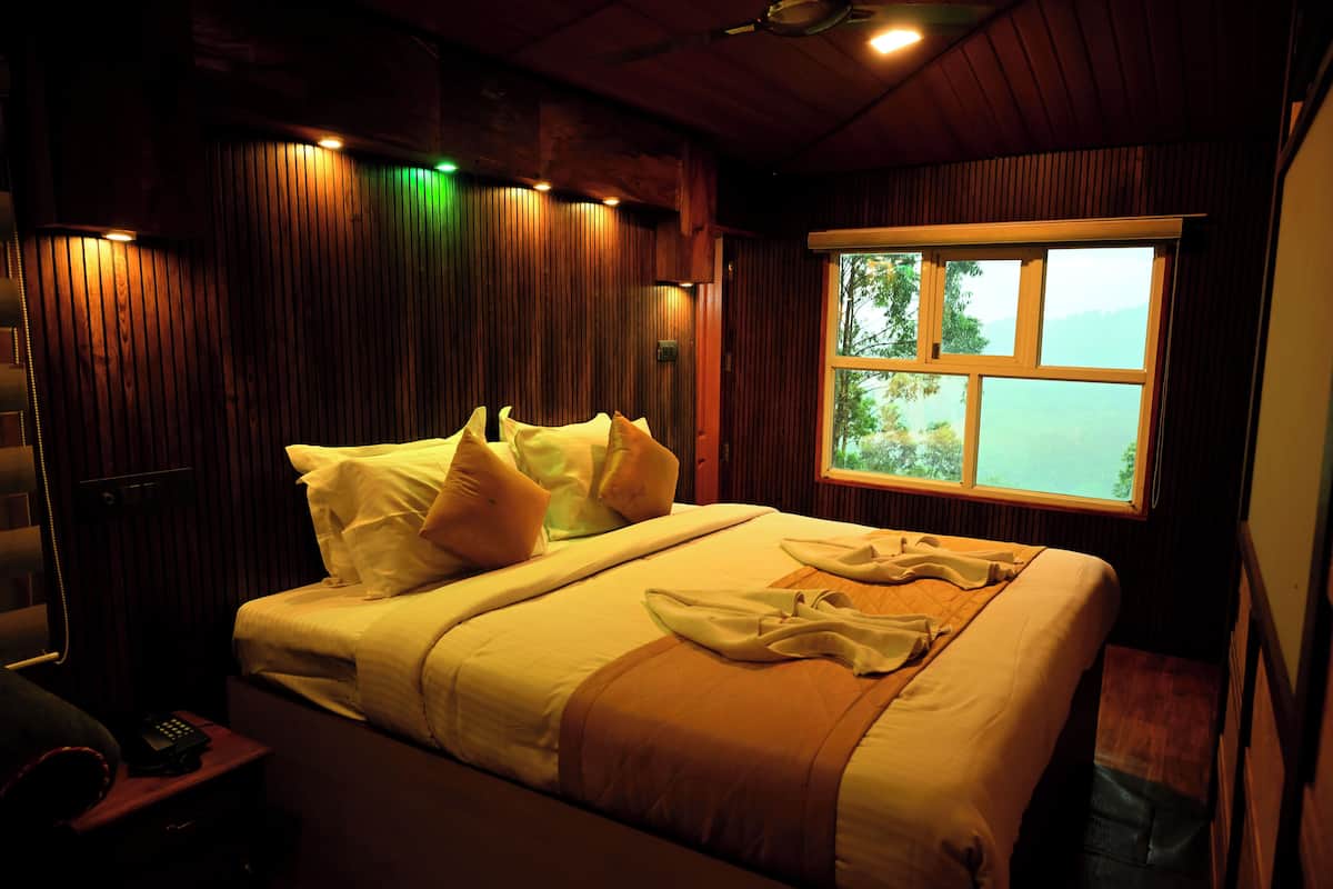 Uma cama bem arrumada com travesseiros e um edredom, uma mesa de cabeceira com um telefone, e uma janela com vista para o exterior.