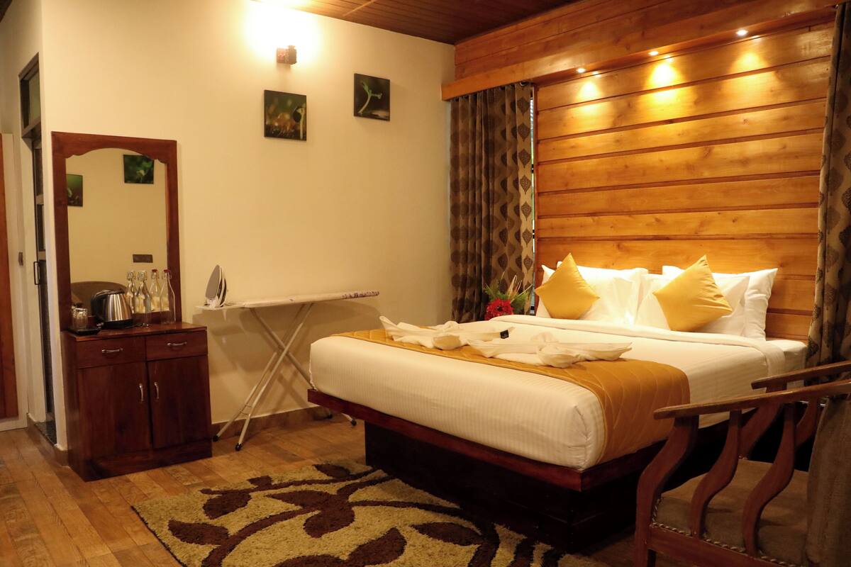 Um quarto com cabeceira de madeira, cama com roupa de cama branca e almofadas amarelas, penteadeira de madeira, espelho e uma pequena mesa com chaleira.