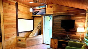 A wooden cabin interior with a glass table, a green sofa, a TV, and a wooden bench.
