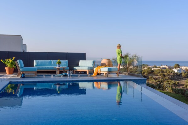 Ein Poolbereich mit einer Sitzecke, einer Person mit Hut und Blick aufs Meer.