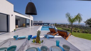 Ett modernt utomhusområde för måltider med en pool och utsikt över havet, där ett bord är dukat med blå tallrikar och en ananas som mittpunkt på bordet.