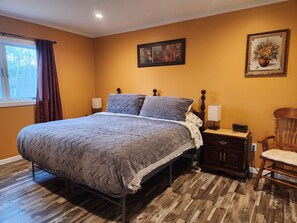 Un dormitorio con una cama grande, piso de madera, una mesita de noche con lámpara, una silla y un cuadro en la pared.