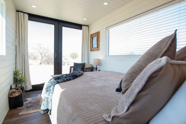 A bedroom with a large bed, a chair, a small table with a lamp, and a window with blinds.