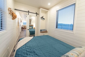 A bedroom with a bed, a window with blinds, a door leading to another room, and a wall-mounted rack. - New! Southwest Shipping Container-In Alpine (Alpine)