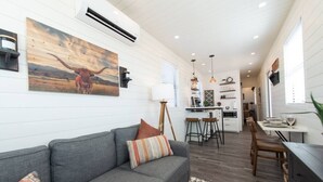 Un salon moderne avec un canapé gris, un coin repas avec une petite table et des chaises, et une cuisine en arrière-plan.