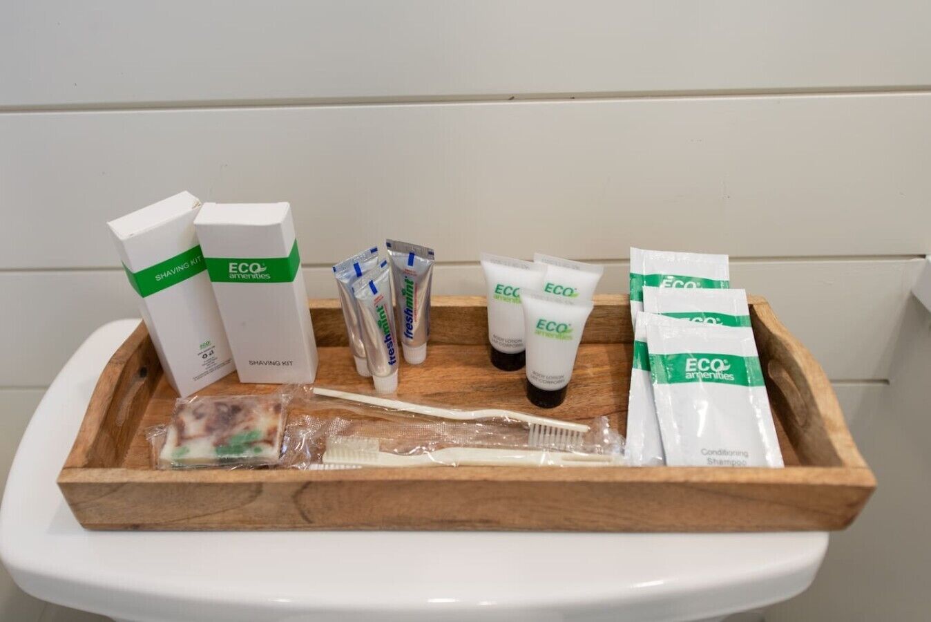 Un plateau en bois contenant un kit de rasage, un shampooing à la menthe fraîche et un après-shampooing, ainsi qu’une brosse à dents, posés sur les toilettes, qui sont de couleur blanche.