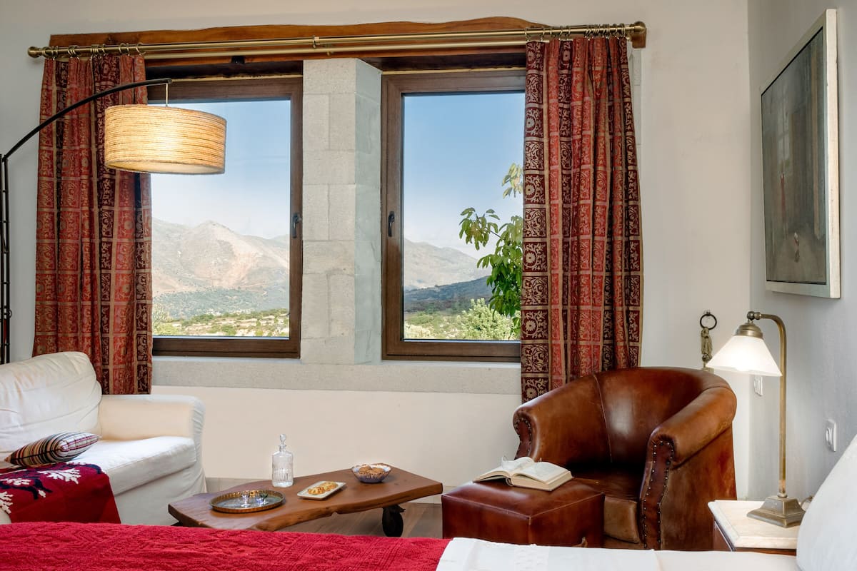 Una habitación de hotel con un ventanal amplio, un sofá, una silla, una mesa con bandeja y una lámpara.