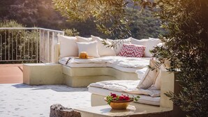 A cozy outdoor seating area with a cushioned bench, a bowl of fruit, and a view of a mountainous landscape. - Nassibe (Mylopotamos)