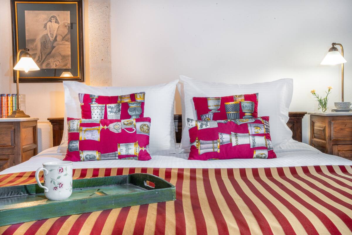 Una cama bien hecha con almohadas estampadas, una mesita de noche con una lámpara y un cuadro en la pared.