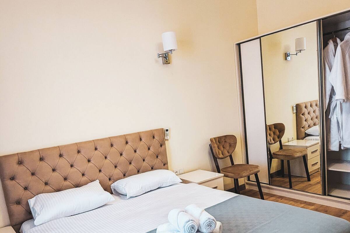 A hotel room with a large bed, a bedside table, a chair, a mirror, and a wardrobe.