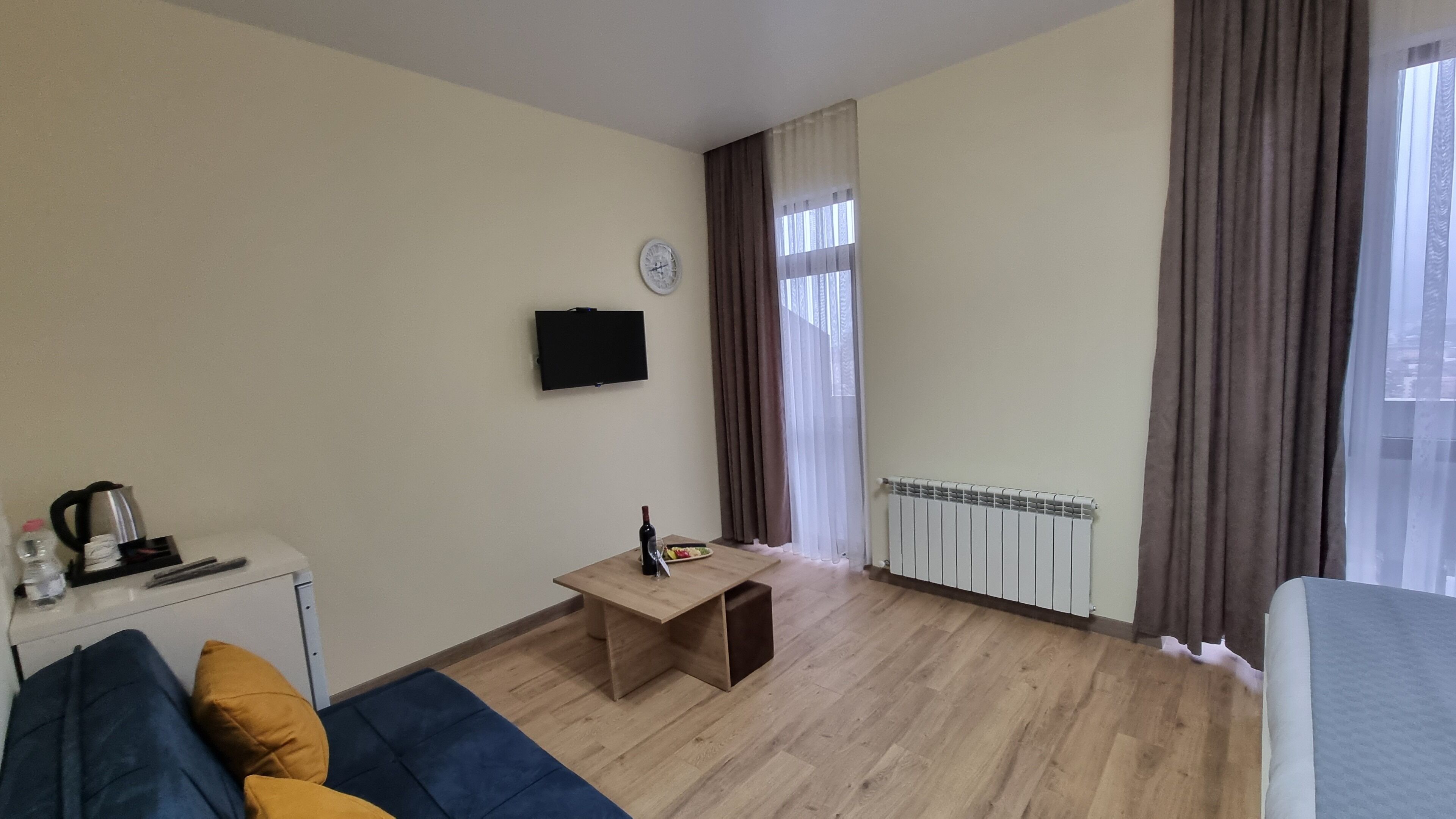A hotel room with a wooden floor, a bed, a sofa, a small table with a bottle and glass, a kettle, a TV on the wall, and a window with curtains.