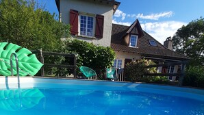 A swimming pool with a wooden deck in the foreground, a large inflatable leaf decoration, and a house with a red window shutter in the background. - Beautiful Quercy house near the Chemin de St Jacques (LUNAN)