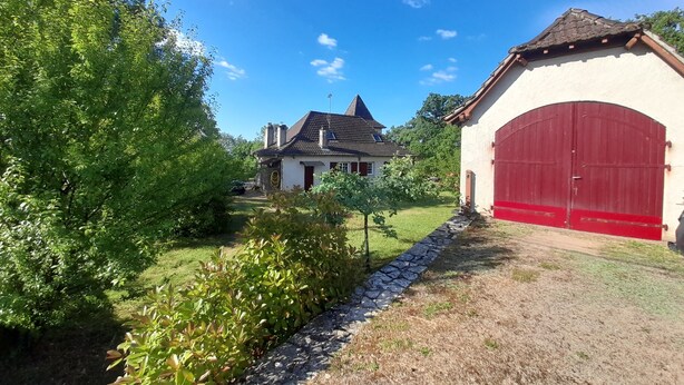 A house with a red garage door, a sloped roof, and a small garden area. - Beautiful Quercy house near the Chemin de St Jacques (LUNAN)