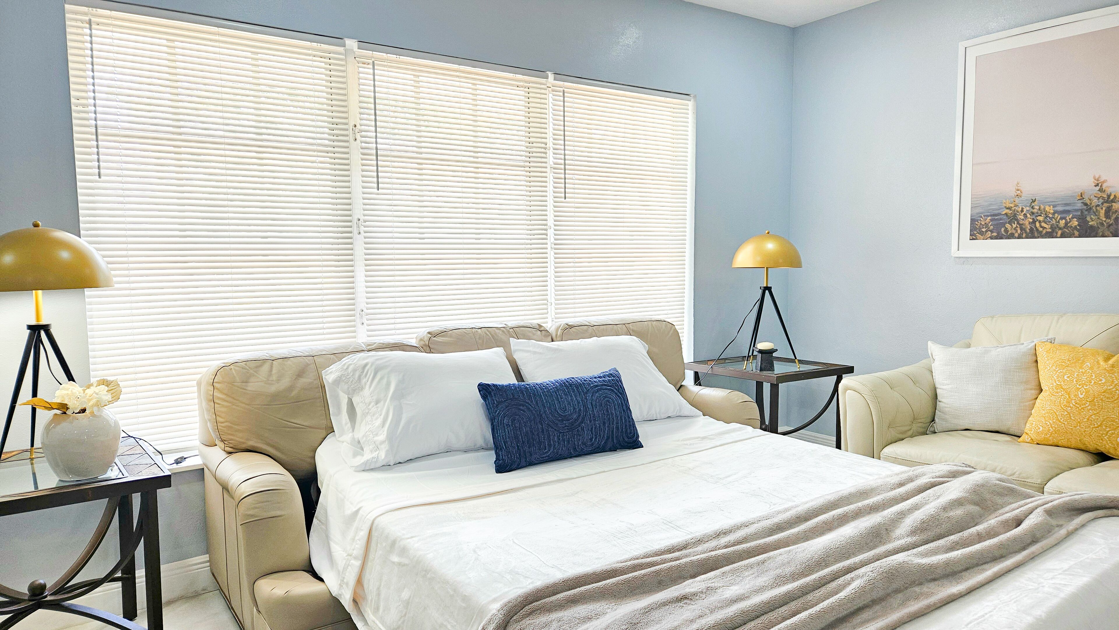 A bedroom with a bed, two bedside tables, a lamp, a chair, and a window with blinds.