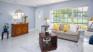 A spacious living room with a beige sofa, a wooden cabinet, a coffee table, and a large window offering a view of greenery. - Paradise Vacation Home for entire family (Boynton Beach)
